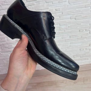 May include: A black leather lace-up shoe featuring a smooth, polished finish and a thick, black sole. The midsole is accented with a textured, grey, snakeskin-patterned trim that wraps around the entire perimeter of the shoe above the outsole.