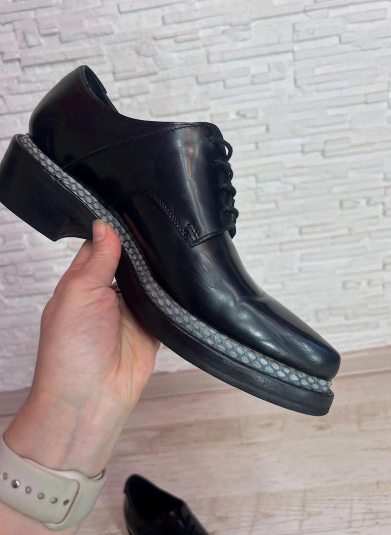May include: A black leather lace-up shoe featuring a smooth, polished finish and a thick, black sole. The midsole is accented with a textured, grey, snakeskin-patterned trim that wraps around the entire perimeter of the shoe above the outsole.