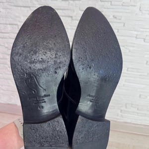 May include: A pair of black shoes with pointed toes and low heels. The soles are black and feature the embossed text Acne Studios, Made in Italy, and the size 40. The soles show signs of wear with scuffs and texture across the surface.
