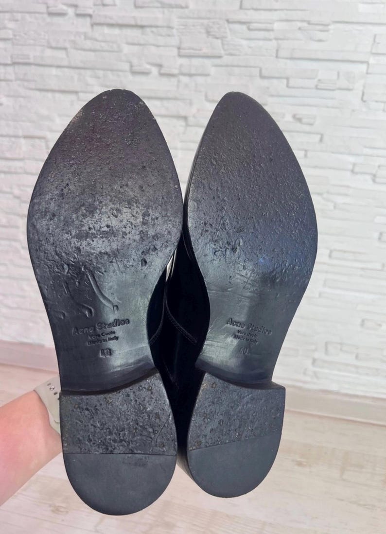 May include: A pair of black shoes with pointed toes and low heels. The soles are black and feature the embossed text Acne Studios, Made in Italy, and the size 40. The soles show signs of wear with scuffs and texture across the surface.