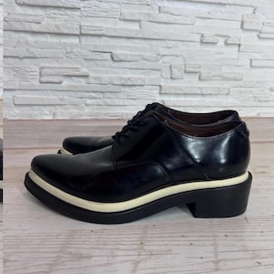 May include: A pair of black leather lace-up shoes featuring a pointed toe, a thick white midsole, and a solid black block heel. The shoes have a polished finish, black laces, and a low-cut ankle profile with a structured, smooth leather upper design.
