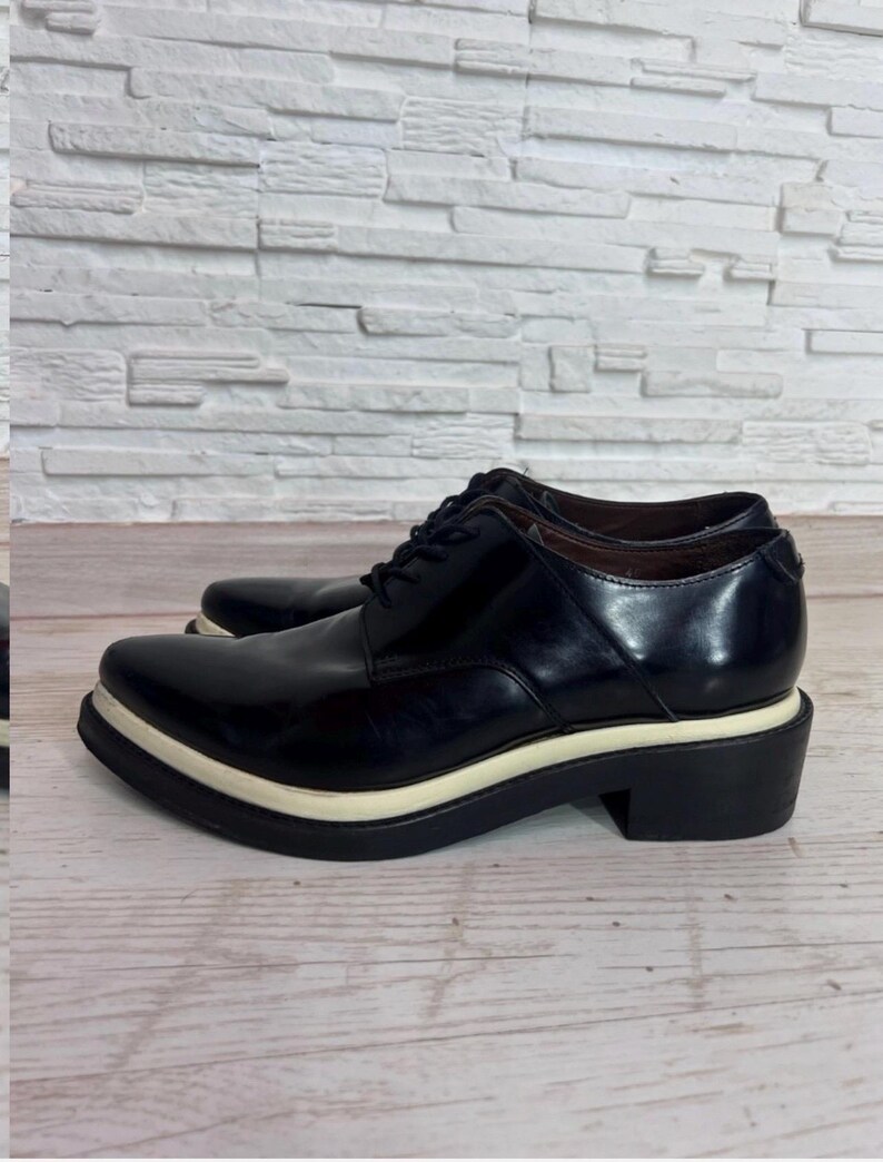 May include: A pair of black leather lace-up shoes featuring a pointed toe, a thick white midsole, and a solid black block heel. The shoes have a polished finish, black laces, and a low-cut ankle profile with a structured, smooth leather upper design.