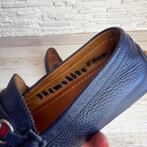 May include: Close-up of a navy blue leather loafer with a silver buckle and red and green accents. The interior is tan leather with stamped text. The shoe is on a light wood surface.