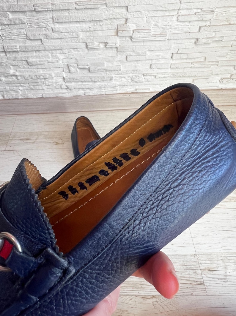 May include: Close-up of a navy blue leather loafer with a silver buckle and red and green accents. The interior is tan leather with stamped text. The shoe is on a light wood surface.
