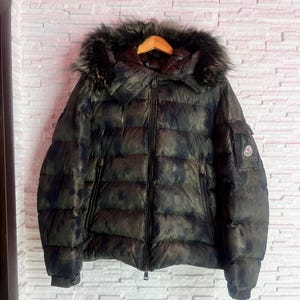 Puede incluir: Una chaqueta acolchada de camuflaje verde oscuro y negro con capucha forrada de piel. La chaqueta tiene una cremallera completa, múltiples bolsillos y un parche con el logotipo en la manga. La chaqueta está colgada en una percha de madera.