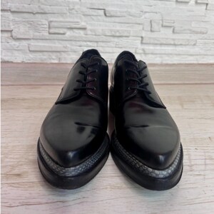 May include: A pair of black leather lace-up shoes with a pointed toe design. The shoes feature a thick black sole with a textured grey snakeskin-patterned trim along the edge of the platform, black laces, and a smooth, polished finish.