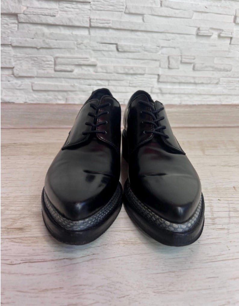 May include: A pair of black leather lace-up shoes with a pointed toe design. The shoes feature a thick black sole with a textured grey snakeskin-patterned trim along the edge of the platform, black laces, and a smooth, polished finish.