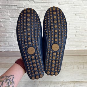 May include: A pair of navy blue leather driving shoes with brown rubber soles. The soles feature a pattern of small, circular grips and the brand name "Gucci" is visible on the sole.