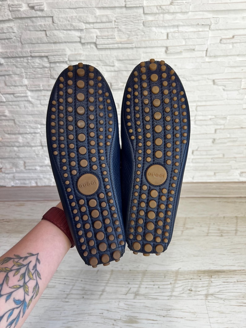 May include: A pair of navy blue leather driving shoes with brown rubber soles. The soles feature a pattern of small, circular grips and the brand name "Gucci" is visible on the sole.
