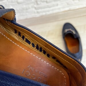 May include: Close-up of a navy blue leather loafer with a brown leather insole. The shoe has a stitched design and the words "made in Italy" are visible. A second loafer is slightly out of focus in the background.