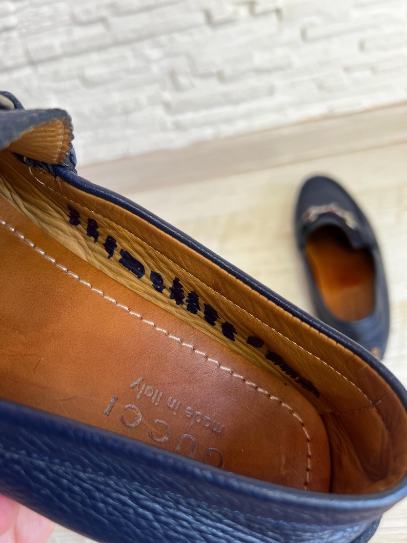 May include: Close-up of a navy blue leather loafer with a brown leather insole. The shoe has a stitched design and the words "made in Italy" are visible. A second loafer is slightly out of focus in the background.