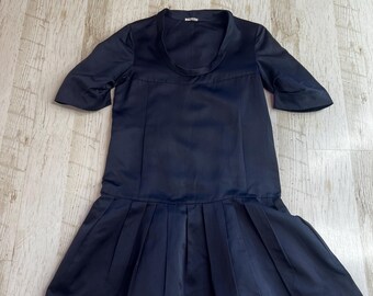 May include: A dark navy blue dress with a smooth, satin-like finish. The garment features a round neckline, short sleeves, and a drop-waist silhouette with a pleated skirt section that flares out from the hips to the hemline.