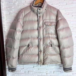 Puede incluir: Una chaqueta acolchada de color gris claro con cierre de cremallera y un bolsillo grande en la parte delantera. La chaqueta tiene un cuello y puños de canalé.