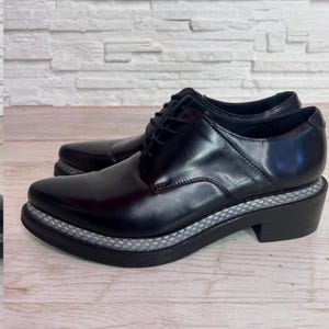 May include: A pair of black leather oxford shoes featuring a pointed toe, black laces, and a thick sole with a textured grey and white snakeskin-patterned trim along the midsole. The shoes have a low block heel and a smooth, polished finish.