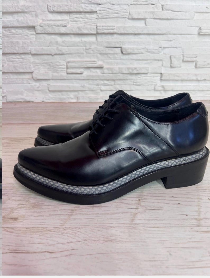 May include: A pair of black leather oxford shoes featuring a pointed toe, black laces, and a thick sole with a textured grey and white snakeskin-patterned trim along the midsole. The shoes have a low block heel and a smooth, polished finish.