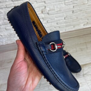 May include: Navy blue leather loafer with a silver buckle and a red and navy stripe detail. The shoe has a textured leather upper and a rubber sole. The word "Aquila" is visible on the inner lining of the shoe.