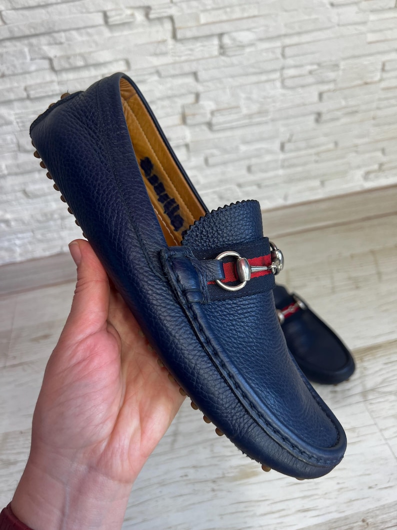 May include: Navy blue leather loafer with a silver buckle and a red and navy stripe detail. The shoe has a textured leather upper and a rubber sole. The word "Aquila" is visible on the inner lining of the shoe.