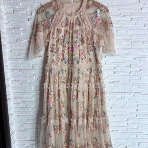 Puede incluir: Un vestido beige hasta la rodilla con una capa de tul transparente y bordados florales multicolores en tonos rosa, amarillo y azul. La prenda presenta mangas cortas con volantes, un cuello alto y un diseño de falda con niveles.
