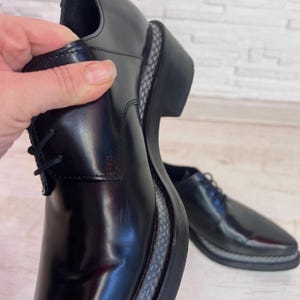 May include: A pair of black leather lace-up shoes with a polished finish, featuring a thick black sole with a grey and black patterned textured welt detail along the edge, and black laces.