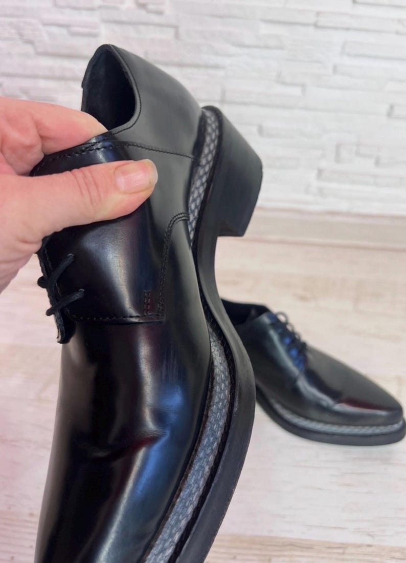 May include: A pair of black leather lace-up shoes with a polished finish, featuring a thick black sole with a grey and black patterned textured welt detail along the edge, and black laces.