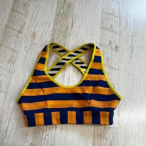 May include: A sports bra featuring horizontal navy blue and orange stripes with bright yellow trim. The back design includes crisscross straps with a black and white diagonal striped pattern. The bottom band features vertical navy blue and orange stripes.