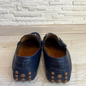 May include: A pair of navy blue leather driving loafers. The shoes have a classic design with a rounded toe, a leather interior, and a rubber sole with small, circular grips. The shoes are displayed against a white and wood background.