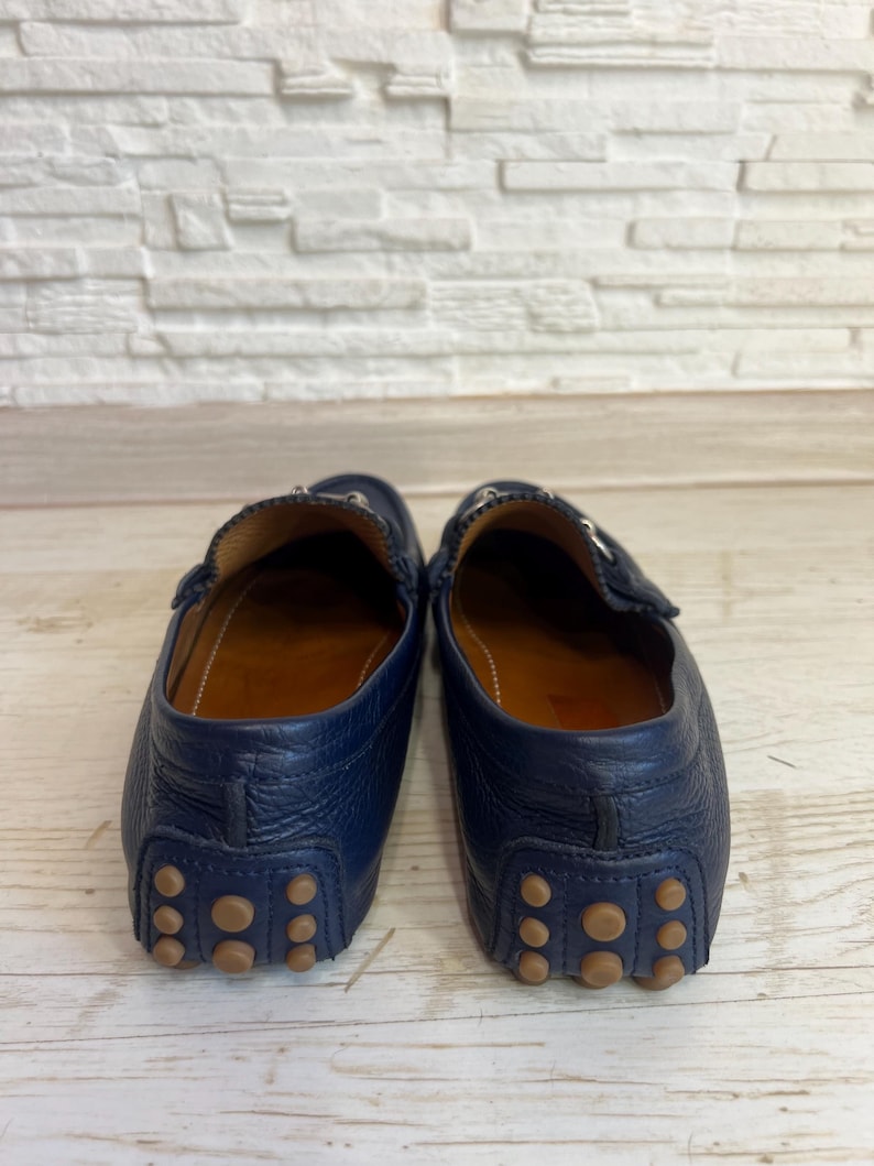 May include: A pair of navy blue leather driving loafers. The shoes have a classic design with a rounded toe, a leather interior, and a rubber sole with small, circular grips. The shoes are displayed against a white and wood background.