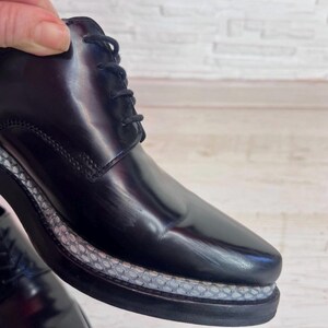 May include: A black lace-up shoe with a polished, glossy finish. The shoe features a thick, dark sole with a textured, light gray snakeskin-patterned trim running along the midsole. The design includes black laces and visible stitching.