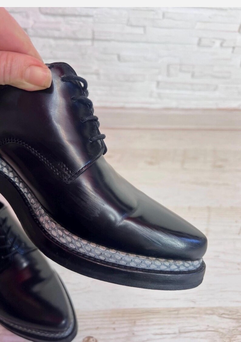 May include: A black lace-up shoe with a polished, glossy finish. The shoe features a thick, dark sole with a textured, light gray snakeskin-patterned trim running along the midsole. The design includes black laces and visible stitching.