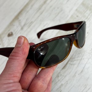 May include: Brown Ray-Ban sunglasses with dark green lenses. The sunglasses have a curved frame and are held in a hand. The Ray-Ban logo is visible on the lens.