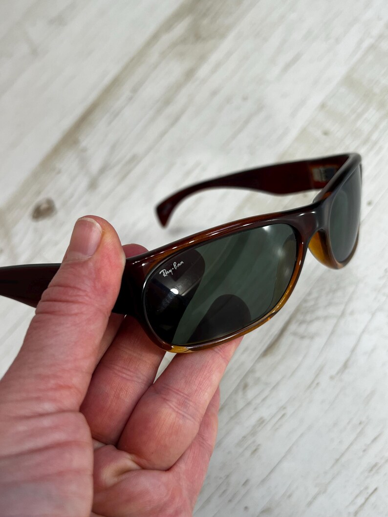 May include: Brown Ray-Ban sunglasses with dark green lenses. The sunglasses have a curved frame and are held in a hand. The Ray-Ban logo is visible on the lens.