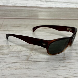 May include: Brown sunglasses with dark green lenses. The sunglasses have a rectangular frame with a slightly curved design. The lenses are dark, and the frame is a solid brown color. The sunglasses are resting on a light-colored wooden surface.