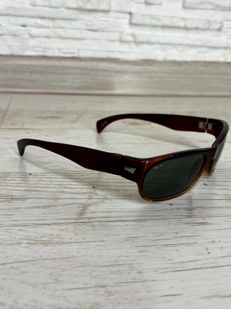May include: Brown sunglasses with dark green lenses. The sunglasses have a rectangular frame with a slightly curved design. The lenses are dark, and the frame is a solid brown color. The sunglasses are resting on a light-colored wooden surface.