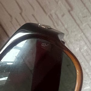 May include: Close-up of sunglasses with a dark brown frame and green lenses. The frame has a tortoiseshell pattern with a gold-colored hinge. The lenses reflect the surrounding environment.
