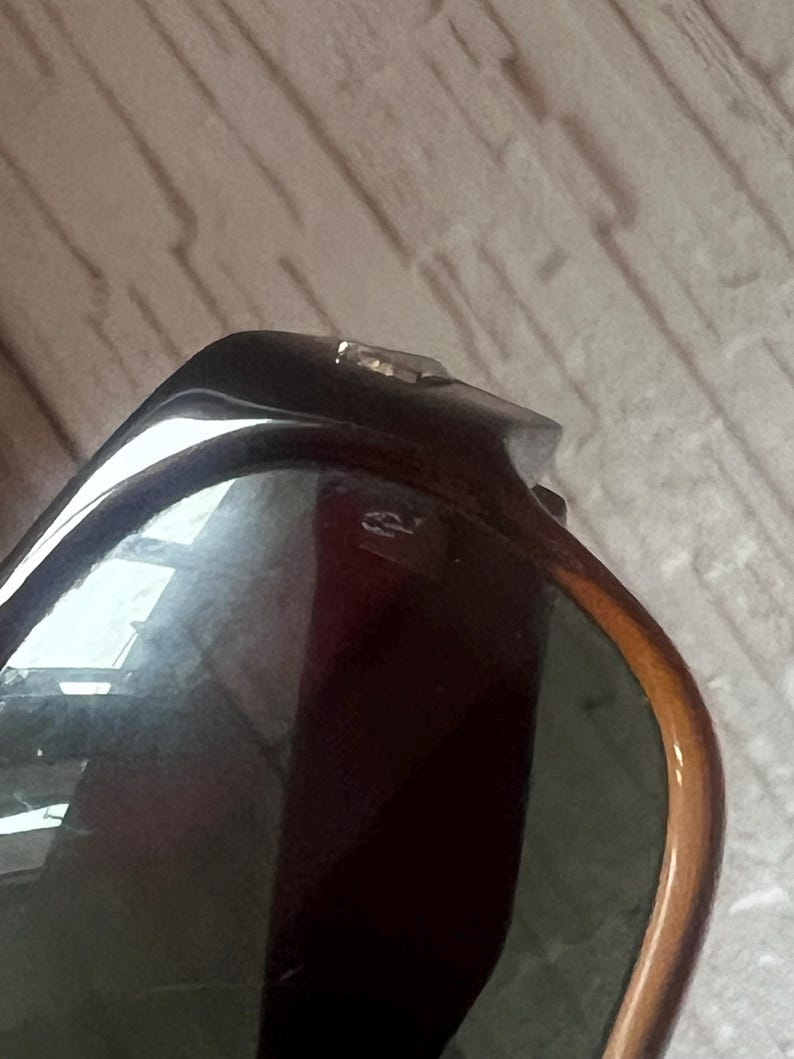 May include: Close-up of sunglasses with a dark brown frame and green lenses. The frame has a tortoiseshell pattern with a gold-colored hinge. The lenses reflect the surrounding environment.