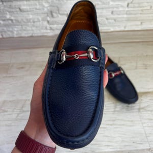 May include: A pair of navy blue leather loafers with a horsebit detail and a red and navy stripe. The shoes are displayed on a light background.