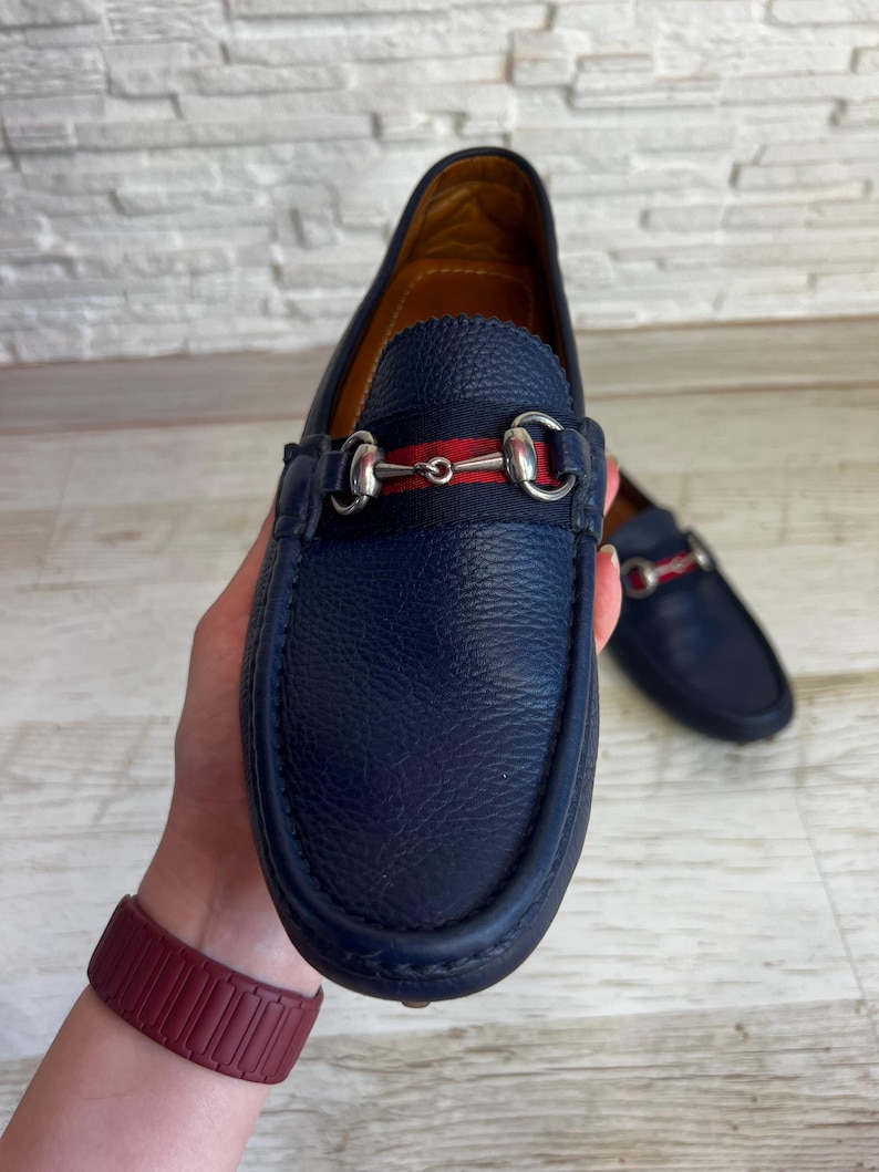 May include: A pair of navy blue leather loafers with a horsebit detail and a red and navy stripe. The shoes are displayed on a light background.