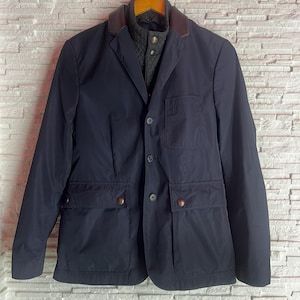 May include: A navy blue jacket with a brown leather collar and a quilted vest underneath. The jacket has two front pockets with button closures and a three-button closure. The jacket is hanging on a wooden hanger against a white brick wall.