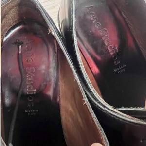 May include: A pair of black leather shoes with a dark interior lining. The inner sole features the text Acne Studios and Made in Italy printed in silver. A thin black lace is visible extending from the interior of one of the shoes.