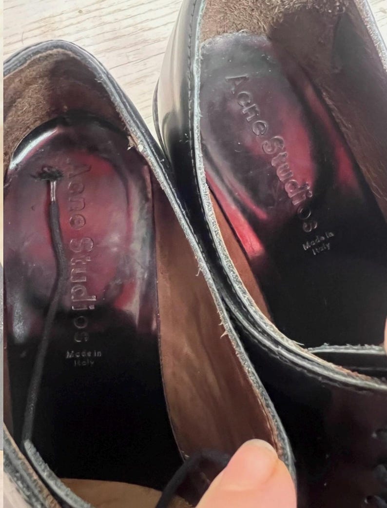May include: A pair of black leather shoes with a dark interior lining. The inner sole features the text Acne Studios and Made in Italy printed in silver. A thin black lace is visible extending from the interior of one of the shoes.