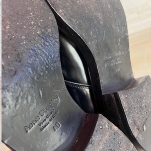 May include: The soles of a pair of black leather shoes with a stacked heel. The soles are dark brown and show signs of wear. Embossed text on the sole reads Acne Studios, Vero Cuoio, Made in Italy, and the size 40.