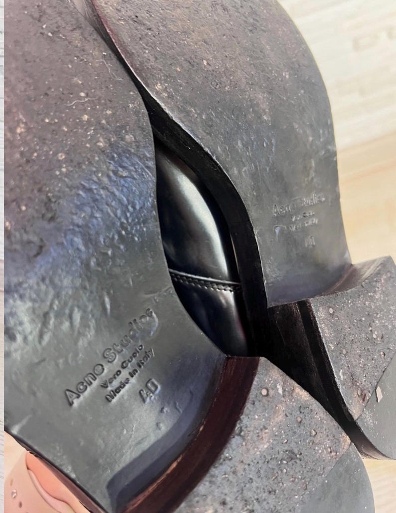 May include: The soles of a pair of black leather shoes with a stacked heel. The soles are dark brown and show signs of wear. Embossed text on the sole reads Acne Studios, Vero Cuoio, Made in Italy, and the size 40.