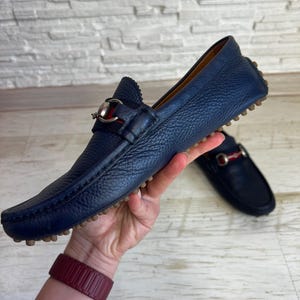 May include: A pair of navy blue leather loafers with a silver and red accent. The shoes have a textured finish and a rubber sole. One shoe is in focus, while the other is slightly blurred in the background. The shoes are held up by a hand.