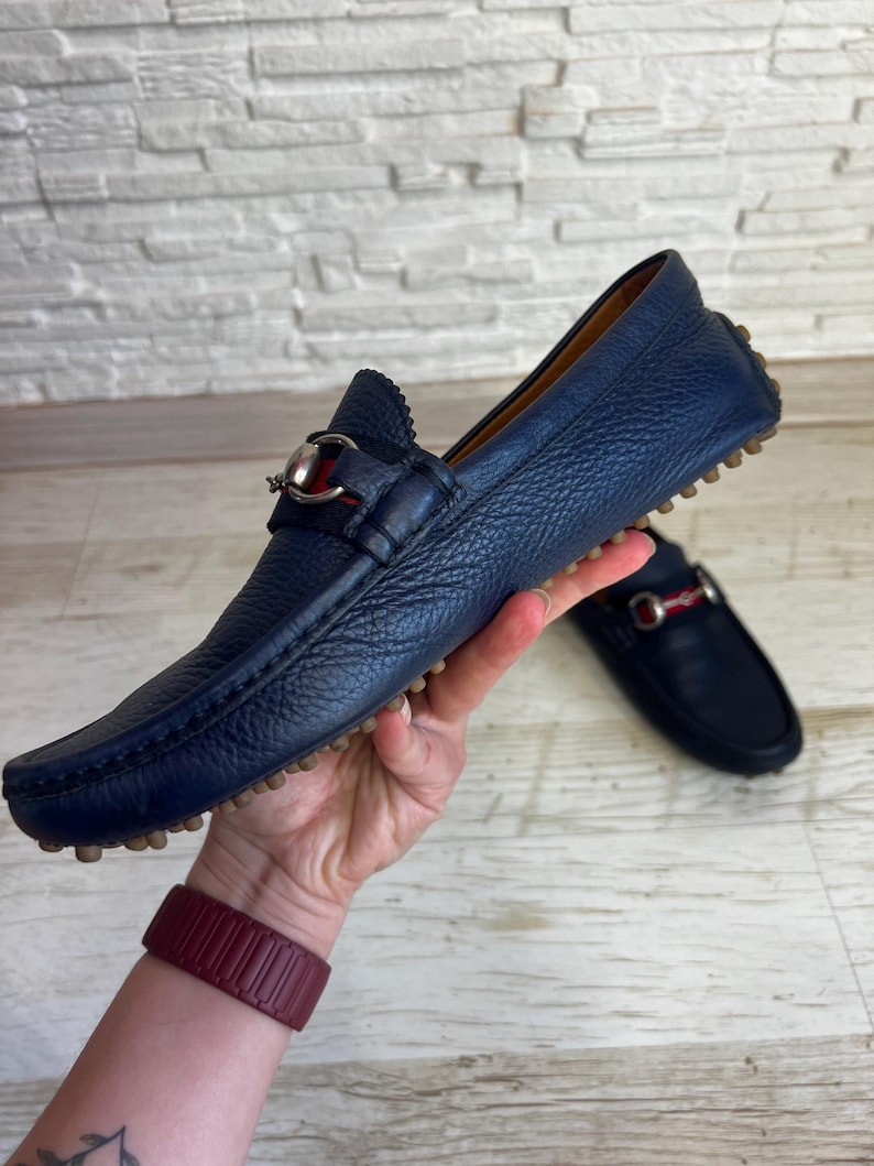 May include: A pair of navy blue leather loafers with a silver and red accent. The shoes have a textured finish and a rubber sole. One shoe is in focus, while the other is slightly blurred in the background. The shoes are held up by a hand.