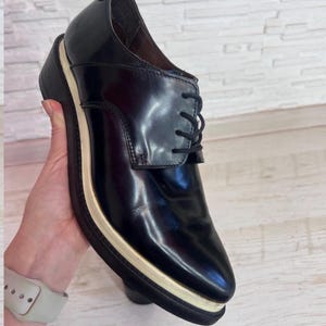 May include: A black leather oxford shoe featuring a polished finish, black laces, and a thick platform sole with a distinct cream-colored midsole layer. The shoe has a rounded toe and a structured silhouette, showcasing a clean design with visible stitching.