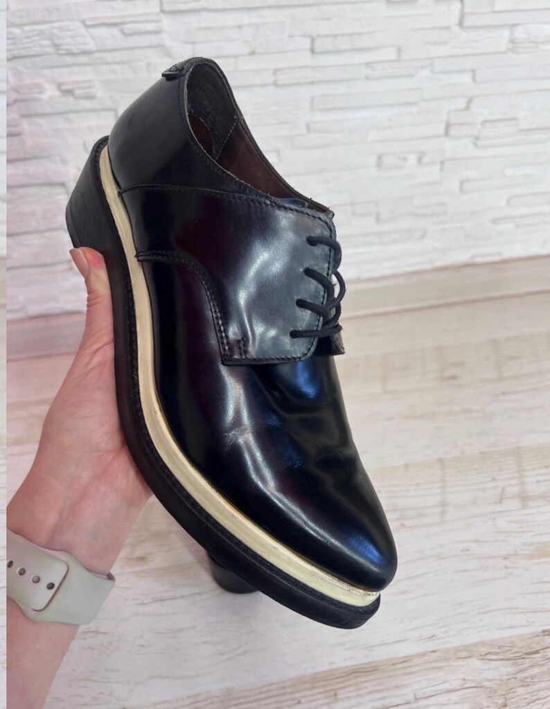 May include: A black leather oxford shoe featuring a polished finish, black laces, and a thick platform sole with a distinct cream-colored midsole layer. The shoe has a rounded toe and a structured silhouette, showcasing a clean design with visible stitching.