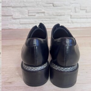 May include: A pair of black leather oxford shoes featuring a smooth, polished finish on the upper and a distinct block heel. The heel base is accented with a textured, grey and white snakeskin-patterned material wrapped around the middle section.