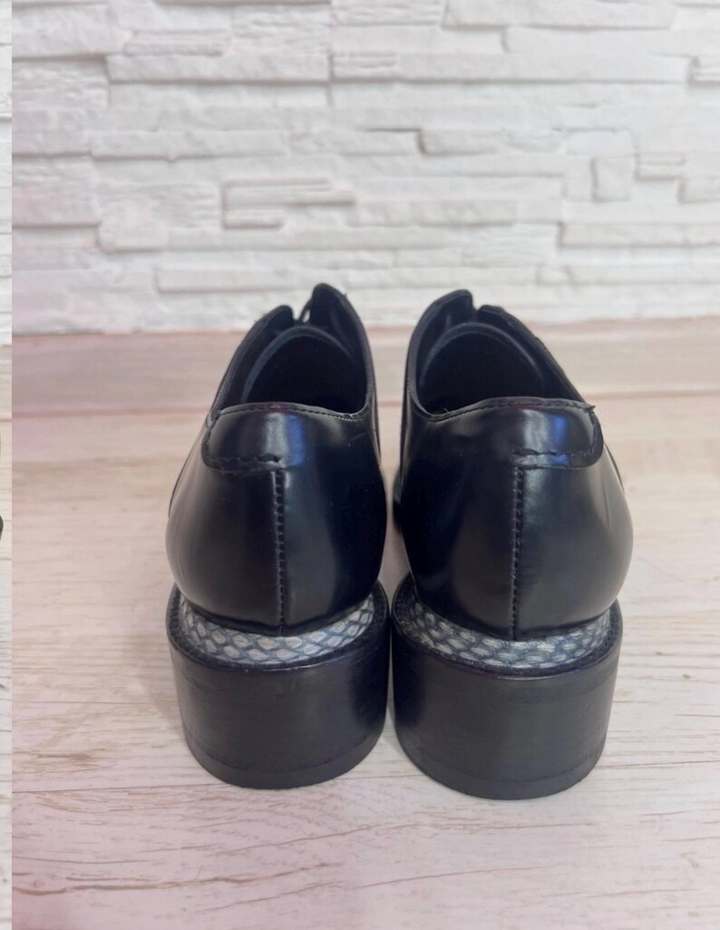 May include: A pair of black leather oxford shoes featuring a smooth, polished finish on the upper and a distinct block heel. The heel base is accented with a textured, grey and white snakeskin-patterned material wrapped around the middle section.