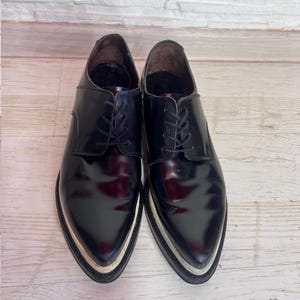 May include: A pair of black leather oxford shoes with a pointed toe, black laces, and a thick, layered white and black sole. The shoes feature a polished, glossy finish and a structured silhouette with a low-cut ankle opening and dark interior lining.