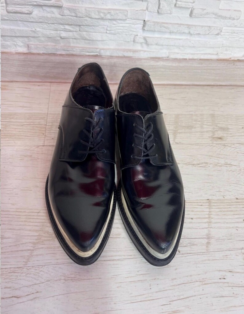 May include: A pair of black leather oxford shoes with a pointed toe, black laces, and a thick, layered white and black sole. The shoes feature a polished, glossy finish and a structured silhouette with a low-cut ankle opening and dark interior lining.